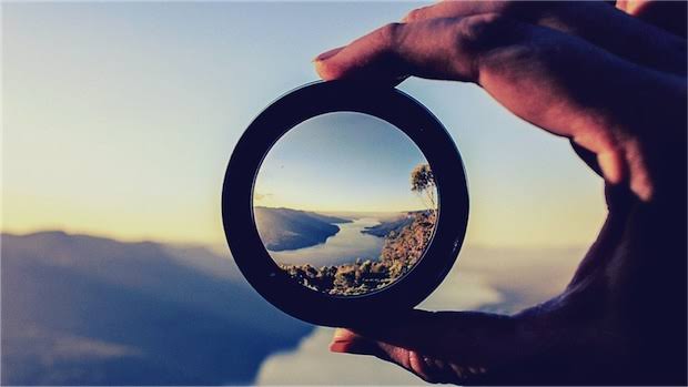 
	A man holds a scop in his right hand while looking in it and over a lake and the sunset 
